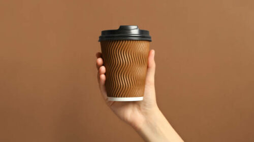 Woman holding takeaway paper coffee cup on brown background, closeup